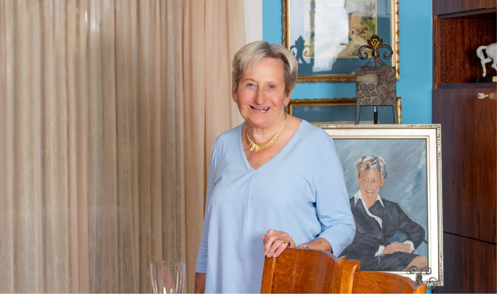 A photo of Margaret Juravinski. She has her hand on the top of a chair and is standing in front of a painted portrait of herself.
